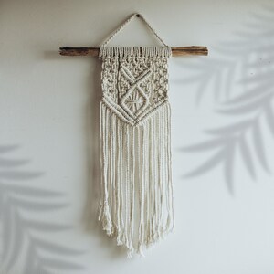 May include: A white macrame wall hanging with a geometric pattern and fringe. The hanging is attached to a wooden dowel.