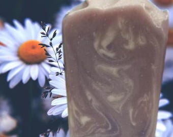 May include: A rectangular bar of handmade soap with a marbled design in shades of brown and cream. The soap has a textured top and is set against a backdrop of white daisies with yellow centers.