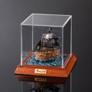 Op de afbeelding: Een miniatuur piratenschip met zwarte zeilen en een schedel en gekruiste beenderen ontwerp wordt getoond in een glazen vitrine op een houten basis. De basis is gelabeld "ElbiLand".