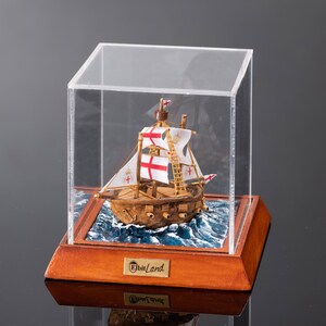 May include: A miniature wooden ship model with white and red sails, displayed in a clear acrylic case on a wooden base. The base has a gold plaque with the text "Elbit Land".