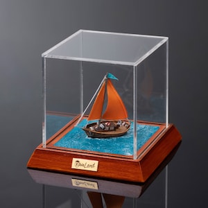 May include: A miniature sailboat with an orange sail and a blue flag is displayed in a clear acrylic case on a wooden base. The base has a gold plaque with the text "ElbiLand" on it.