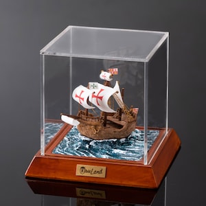 May include: A miniature wooden ship model with white sails and red crosses, displayed in a clear acrylic case on a wooden base. The base has a label that reads "EbiiLand".