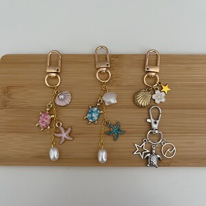 Può includere: Tre portachiavi con chiusure e ciondoli color oro. Uno presenta una tartaruga rosa, una stella marina e una perla. Un altro ha una tartaruga blu, una stella marina e una perla. Il terzo ha una conchiglia, una stella, un fiore, una tartaruga, un'onda e un ciondolo a stella.