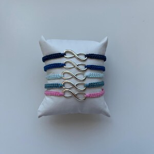 May include: Five friendship bracelets with an infinity symbol charm. The bracelets are made of woven cord in navy blue, light blue, teal, and pink. The infinity charms are silver. The bracelets are displayed on a white pillow.