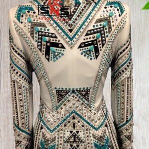 K&ouml;nnte beinhalten: Wei&szlig;e Showjacke mit geometrischem Design mit t&uuml;rkisfarbenen, schwarzen und silbernen Verzierungen. Die Jacke hat einen Stehkragen und lange &Auml;rmel.