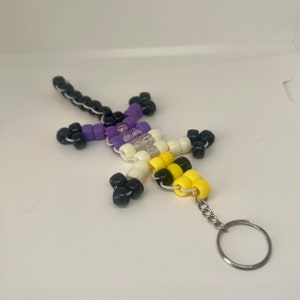 Peut inclure: Un porte-clés en forme de lézard composé de perles violettes, blanches, noires et jaunes. Le porte-clés est doté d'un anneau en métal argenté.