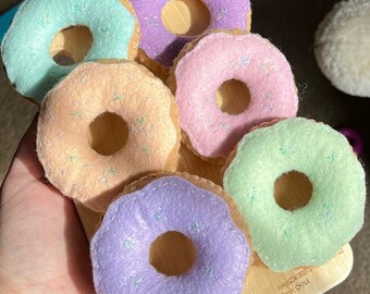May include: Six felt donuts in various pastel colors, including pink, purple, and green. Each donut has a white hole in the center and is decorated with white sprinkles.