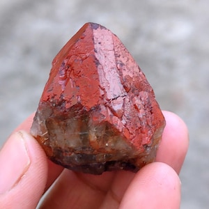 May include: A natural, raw crystal with a deep red hue at the top, transitioning to a lighter, translucent base. The crystal has a rough, textured surface and a pointed, pyramidal shape. The crystal is held in a hand.