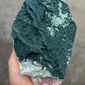 Könnte beinhalten: Ein grobes Mineralexemplar mit einer strukturierten, dunkelblauen Oberfläche. Der Stein hat eine hellere blaugrüne und rosa-weiße Basis. Das Exemplar wird in einer Hand gehalten und zeigt seine natürliche, unregelmäßige Form und Farbvariationen.