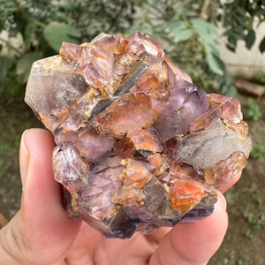 May include: A raw mineral cluster featuring a mix of translucent purple amethyst crystals and opaque, iron-stained orange and brown quartz formations. The specimen displays jagged, multi-faceted surfaces with varying levels of clarity and color saturation.