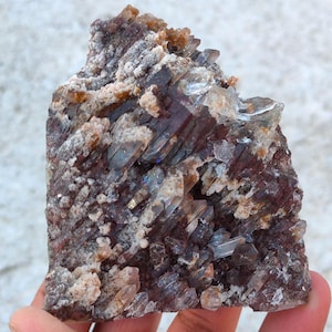 Pode incluir: Um aglomerado de cristais brutos e não cortados com uma mistura de cores. A base é de um tom bordô profundo, com cristais de quartzo claros e esfumaçados. A superfície tem uma aparência áspera e texturizada com manchas bege e brancas.