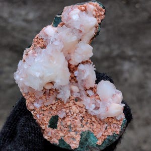 Op de afbeelding: Een natuurlijk mineraal specimen met clusters van witte en lichtroze kristallen op een matrix van roodbruine en groene tinten. Het ruwe, getextureerde oppervlak toont de ruwe schoonheid van de geologische formatie. Een uniek stuk voor verzamelaars.