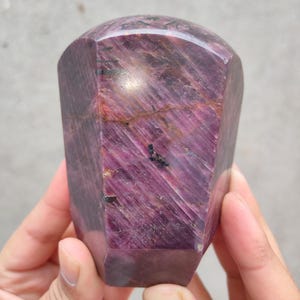 May include: A faceted, deep burgundy crystal with a polished, rounded top. The gemstone has visible striations and dark inclusions, held in a person's hands. The crystal's shape is a truncated hexagonal prism.