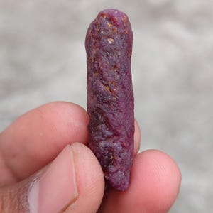 Könnte beinhalten: Ein roher, ungeschliffener Rubin-Edelstein. Der Rubin hat eine tiefe, rot-violette Farbe mit einer rauen, strukturierten Oberfläche. Der Edelstein wird in einer Hand vor einem unscharfen Hintergrund gehalten.
