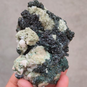 May include: A natural mineral specimen featuring a mix of dark green, black, and light green crystalline structures. The mineral has a rough, textured surface with patches of white and pink hues.