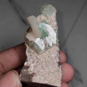 Könnte beinhalten: Mineralprobe mit hellgrünen Kristallen auf einer beige-braunen Gesteinsmatrix. Die Kristalle sind durchscheinend, und das Gestein hat eine raue, strukturierte Oberfläche. Die Probe wird in einer Hand gehalten.