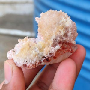 May include: A raw mineral specimen featuring a cluster of pale pink and cream-colored crystalline formations. The surface has a textured, spiky appearance with patches of deep reddish-brown coloration concentrated along the lower edges.