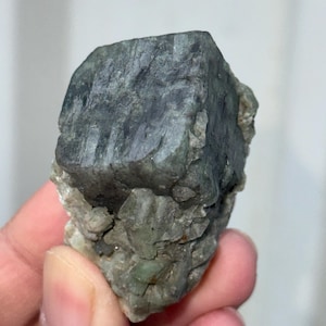 Könnte beinhalten: Ein rohes, ungeschnittenes Mineralexemplar mit einer dunkelgrauen, kristallinen Struktur. Der Stein hat eine raue, strukturierte Oberfläche und eine hellere, grüngraue Basis. Das Mineral wird in einer Hand gehalten und zeigt seine natürliche Form und Farbvariationen.
