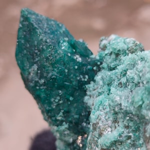 May include: A raw mineral specimen featuring a prominent, deep emerald green crystalline point with a textured, faceted surface. Adjacent to the main crystal is a cluster of smaller, lighter mint green mineral formations with a granular appearance.