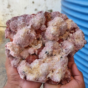 Könnte beinhalten: Ein rohes Mineralexemplar mit einer Ansammlung faseriger, nadelartiger Kristallformationen in den Farben Altrosa, Dunkelrot und Creme. Die Oberfläche hat ein strukturiertes, traubiges Aussehen mit kleinen dunklen Einschlüssen.
