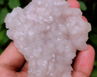 May include: A light pink, crystalline mineral cluster with sharp, pointed formations. The mineral is held in a hand, with a blurred green background. The crystals have a slightly translucent appearance.