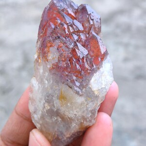 May include: A natural crystal cluster with a mix of clear, reddish-brown, and white colours. The crystal has a rough, textured surface and a cluster of pointed formations. The crystal is held in a hand.