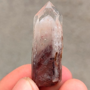 May include: A clear quartz crystal point with internal red and white inclusions. The crystal is held in a hand, showcasing its natural, unpolished form. The tip is transparent, and the base has a reddish hue.