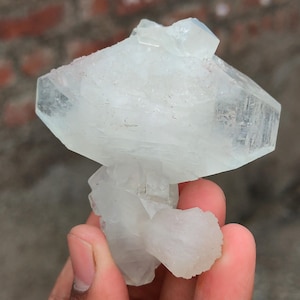 Könnte beinhalten: Ein klarer, kristalliner Mineralienrohling mit einer komplexen, geschichteten Struktur. Der hellfarbene Kristall hat eine geometrische Form mit mehreren Facetten und einer rauen, strukturierten Oberfläche. Das Mineral wird in einer Hand gehalten.
