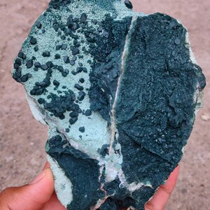 Könnte beinhalten: Nahaufnahme einer Hand, die ein natürliches Mineralexemplar hält. Der Stein weist eine Mischung aus Texturen und Farben auf, mit Bereichen in tiefem Teal, Dunkelgrün und Hellblaugrün. Kleine, dunkle Kristalle sind über die Oberfläche verstreut.