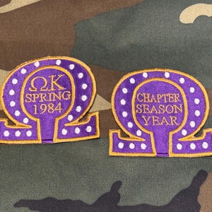 May include: Two purple embroidered patches shaped like the Greek letter Omega, featuring gold borders and white polka dots. The left patch displays the text ΩK SPRING 1984, while the right patch displays the text CHAPTER SEASON YEAR in gold lettering.
