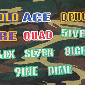 Pode incluir: Uma coleção de patches de tecido com várias palavras e números em fontes arrojadas e coloridas. As palavras incluem "SOLO", "ACE", "DEUCE", "TRE", "QUAD", "5IVE", "6IX", "SE7EN", "8ICHT", "9INE" e "DIME". O fundo é um padrão de camuflagem.
