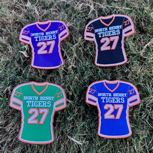 May include: Four jersey-shaped patches in purple, black, green, and blue. Each features the text NORTH HENRY TIGERS, the number 27 on the chest and shoulders, and orange trim. The designs are identical in layout, differing only in the base color.