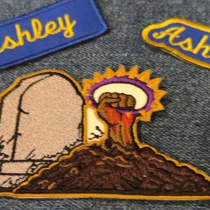 May include: Embroidered patches on denim including two rectangular blue patches with the yellow text Ashley, a patch featuring a brown fist rising from a mound of dirt next to a gray tombstone with a yellow sunburst, and a partial orange patch.