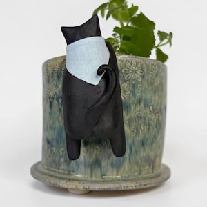 Op de afbeelding: Een cilindervormige keramische plantenpot met een blauw en groen gevlekt glazuur en een reliëf bloemenpatroon. Een zwart kattenfiguurtje met een lichtblauwe driehoekige bandana is aan de voorzijde bevestigd, met zicht op rug, staart en poten.