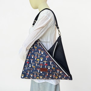 May include: A triangular shoulder bag featuring a dark blue fabric panel with a repeating pattern of various mushrooms in shades of red, yellow, and brown. The bag includes a solid black side panel, a black shoulder strap, and a brown rectangular label.
