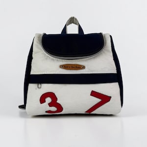 May include: A backpack featuring a white and navy blue color scheme with a top handle and a zippered flap. The front displays the red numbers 3 and 7, and a small brown leather-like label with the text Such-n-Sew Studio is centered on the front.