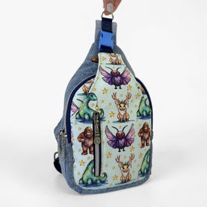 May include: A denim sling backpack featuring a light blue front panel with a repeating pattern of mythical creatures including winged insects, antlered rabbits, and green monsters, accented by yellow stars and a silver vertical zipper.