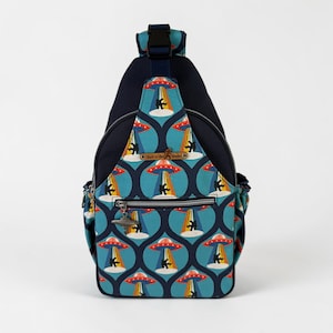 May include: A navy blue and teal sling backpack featuring a repeating pattern of UFOs and silhouettes of Bigfoot. The bag includes a front zippered pocket with a silver-toned UFO zipper pull, a black buckle strap, and a label reading Sew-n-Sew Studio.
