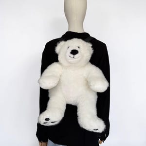 May include: A white plush teddy bear backpack with black accents. The bear has a smiling face, black eyes, and black paw pads. The backpack is worn on a black shirt.