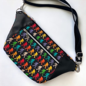 May include: A black fanny pack with a rainbow print of Bigfoot figures. The bag has a black adjustable strap and two zippered compartments.