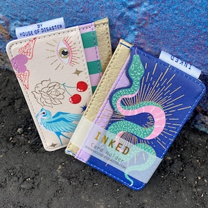 May include: Two card holders with distinctive designs. One has an eye, cherries, and a bird, the other a snake. Both have gold accents and the text "INKED" and "Card Holder". The brand name "HOUSE OF DISASTER" is also visible.