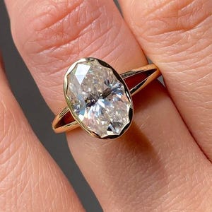 Pode incluir: Um anel de noivado de ouro com um grande diamante de corte oval. O diamante está engastado em uma moldura de bisel e a banda é de ouro polido. O anel é mostrado em uma mão com unhas cuidadas.
