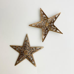 May include: Two ornate, gold-colored star-shaped decorations. Each star features intricate filigree patterns and a central floral design. The stars are displayed against a white background, highlighting their detailed craftsmanship.