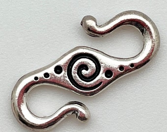 May include: Silver-toned metal clasp with a spiral design and small dots. The clasp is shaped like a figure eight with two loops.