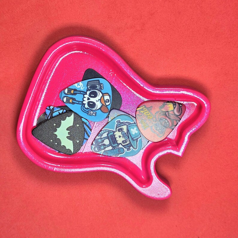 May include: A bright pink guitar-shaped tray containing four guitar picks. The picks feature various designs including a skull with headphones, a bat silhouette on a black background, a robot character, and a textured orange graphic print.