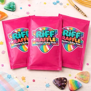 May include: Three bright pink rectangular mystery bags for guitar picks, each featuring a central logo with the text Riff Raffle Mystery Guitar Picks in a colourful rainbow-bordered shape. Several loose, decorative guitar picks are scattered nearby.