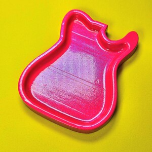 May include: A bright neon pink tray shaped like the body of an electric guitar. The tray features a smooth, glossy finish with a slightly recessed center and raised edges, showcasing a vibrant, solid color throughout its entire guitar-like form.