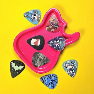 May include: A bright pink guitar-shaped tray holding three guitar picks, including one with a paint can graphic, one with a skull character, and one with a grey marbled pattern. Three additional picks are scattered nearby with skull and bat designs.