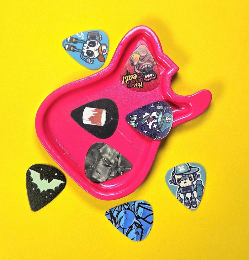 May include: A bright pink guitar-shaped tray holding three guitar picks, including one with a paint can graphic, one with a skull character, and one with a grey marbled pattern. Three additional picks are scattered nearby with skull and bat designs.
