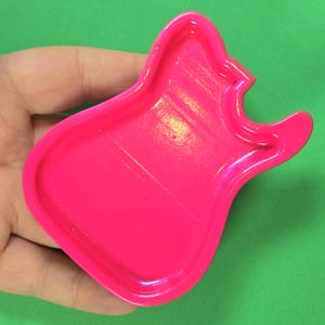 May include: A bright neon pink guitar-shaped cookie cutter with a raised rim and a recessed center. The object features a distinct body shape with a cutout neck and headstock area, designed for cutting dough into the silhouette of an electric guitar.